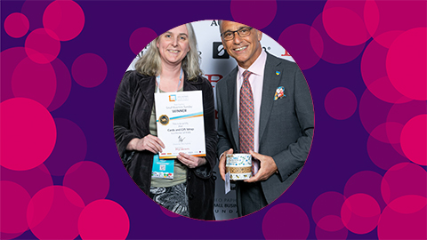 A purple background with pink circles on with an image of Catherine smiling in and holding her certificate next to Theo Paphitis who is holding her tape the middle of the canvas.