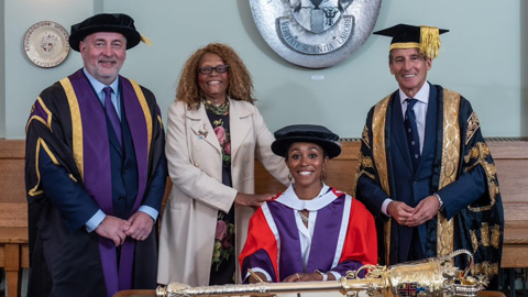 Ebony-Jewel Rainford-Brent is seated at a desk wearing a red robe and hat. Standing to the left is Vice-Chancellor Professor Nick Jennings and her mother, Janet Rainford. On the right is Chancellor, Lord Sebastian Coe. 
