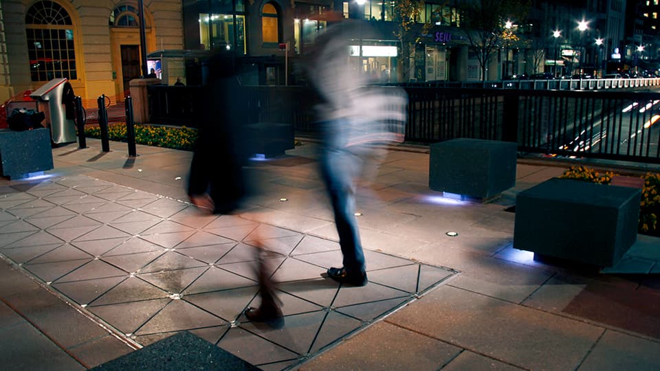 Pavegen tiles in a footpath. There are blurred images of two people walking on the tiles at night time.