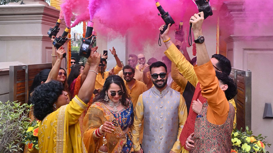 An image of Naami and Suvam on their wedding day with their friends creating a human arch and throwing coloured powder