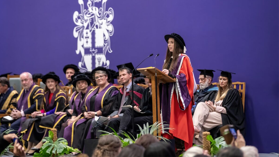Helen standing in front of a podium giving speech at graduation, wearing cap and gown