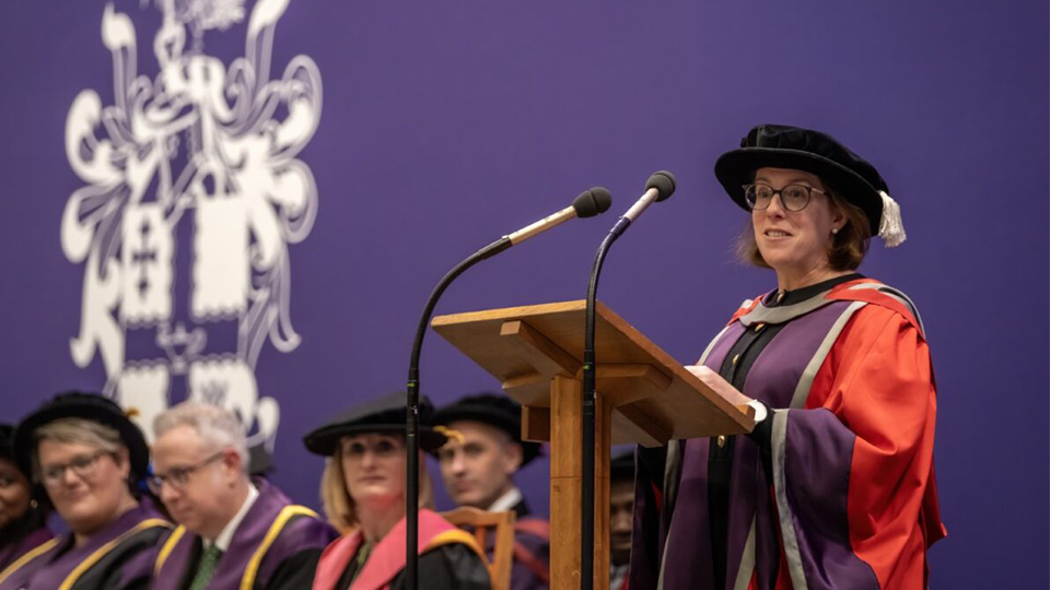 Dr Rebecca standing in front of a podium at graduation wearing cap and gown