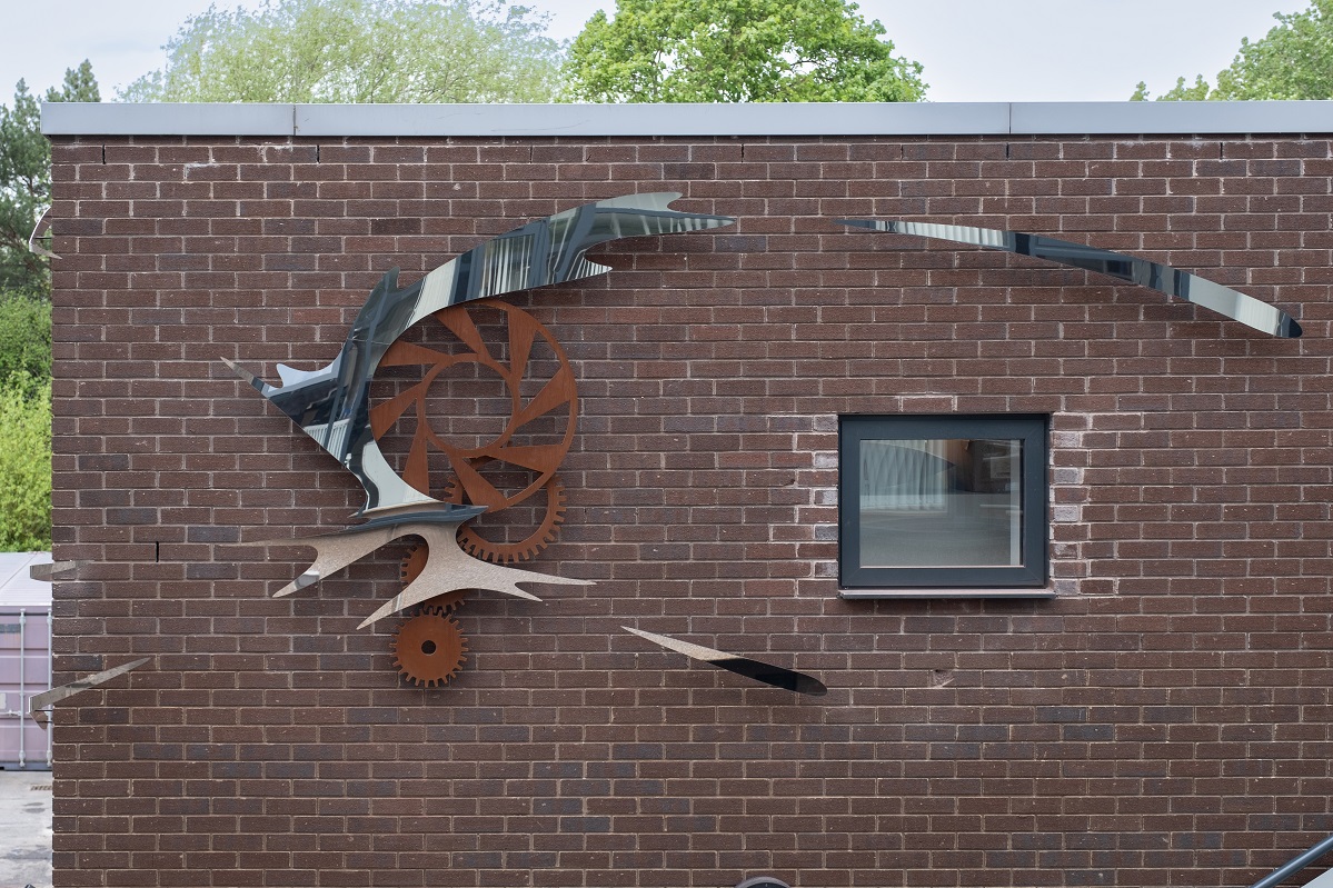 A sculpture attached to the upper corner of a two storey building. It has three corten steel cogs around which a stainless steel form swirls, with separate stainless steel elements giving the impression of being flung off in a circular motion.