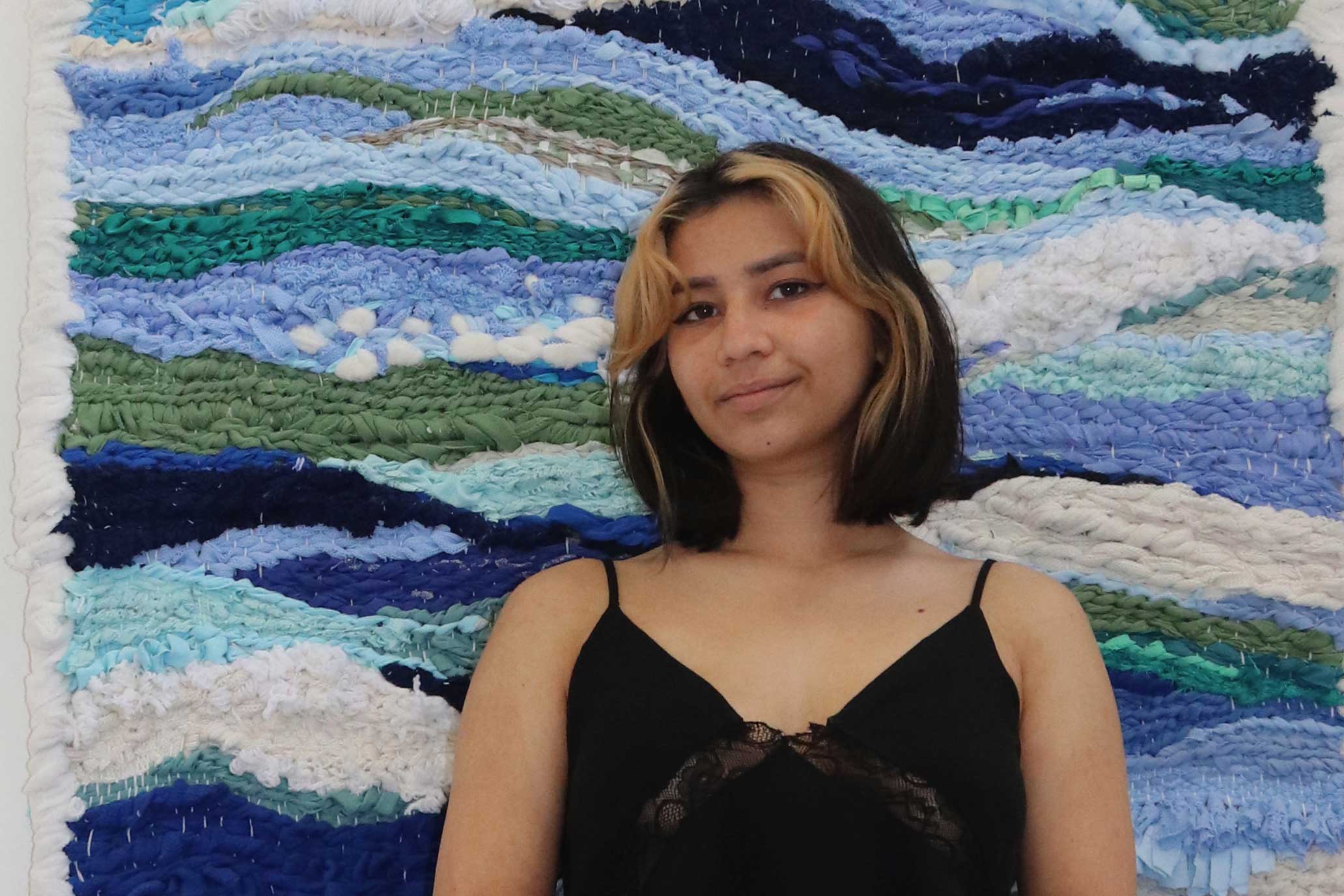 A young, female student is pictured in front of a wall hanging of woven textiles in different shades of blue with some green and white. She has shoulder length dark brown bob with blond streaks at the front and is wearing a black vest top. She is smiling with a closed mouth.