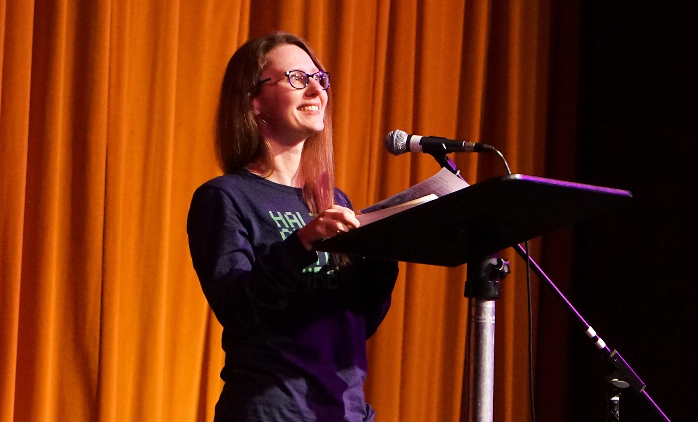 A young white woman in her mid 20s wears glasses and stands in front of a pulpit reading poetry and smiling