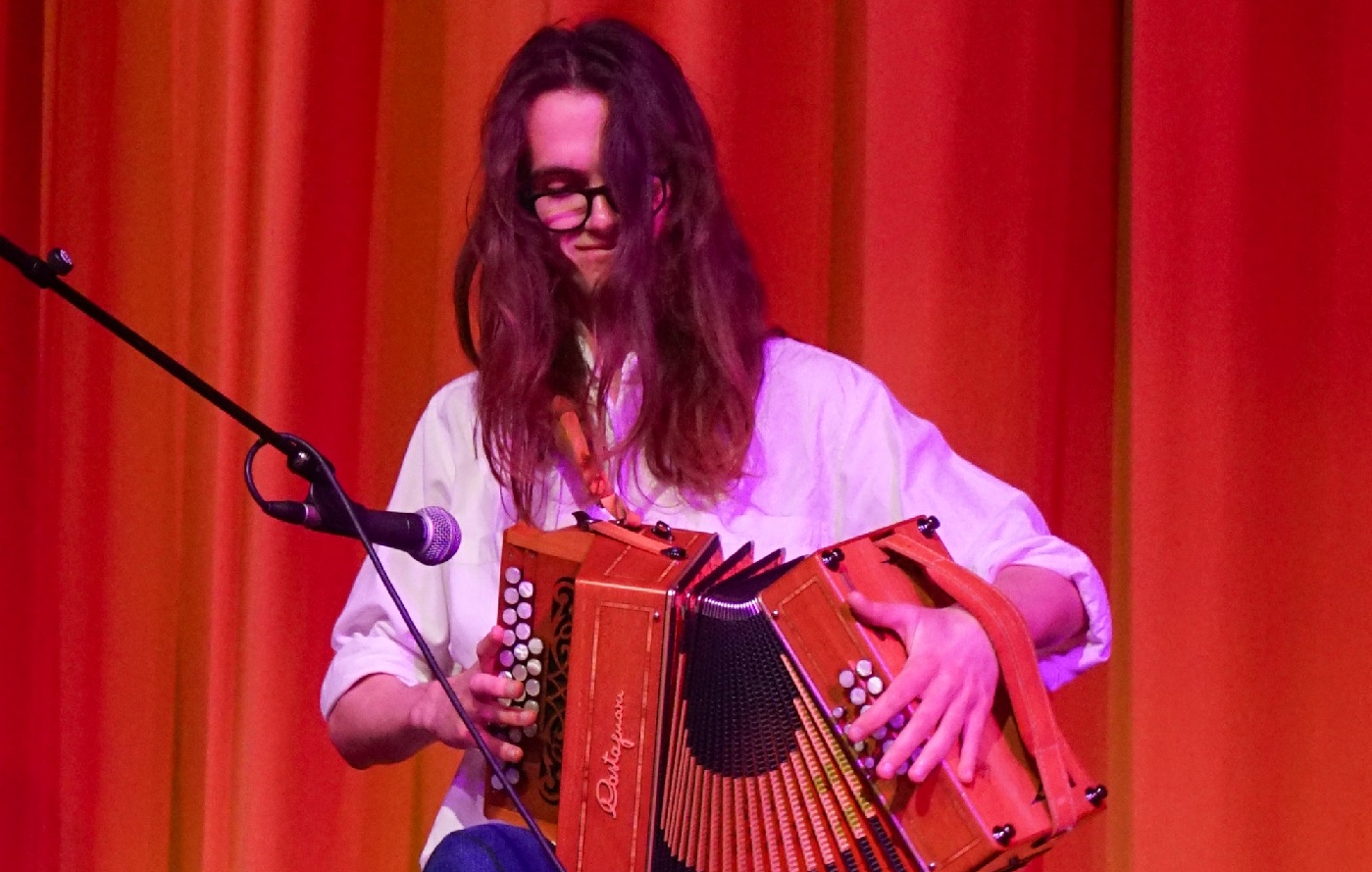 a young man in his early 20s with long brown hair smiling playin an accordion sitting down in front of an orange curtain
