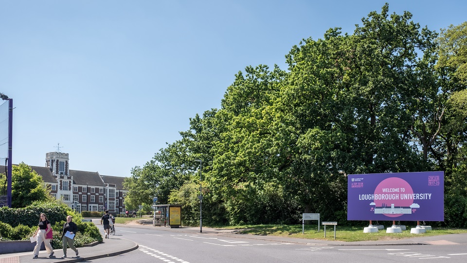 A Welcome To Loughborough University sign flanked by trees in the sunshine. The Hazlerigg building stands in the background.