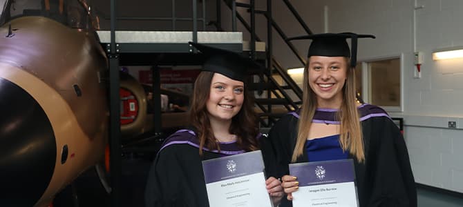 2 female students in gowns and hats holding certificates stood in front of a plane