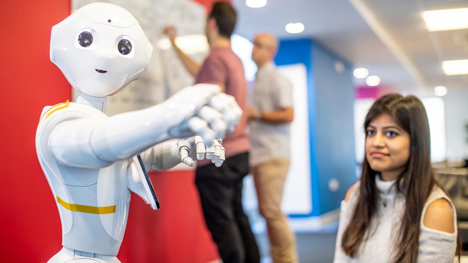 Humanoid robot pepper facing forward with a feamle student slightly blurred in the background.