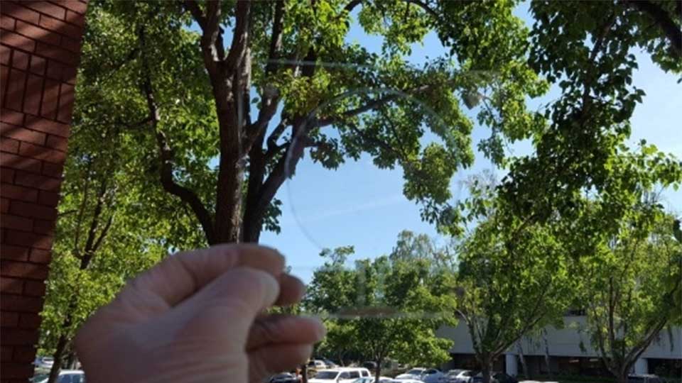 A hand in a thin plastic glove holding a transparent sheet. The plastic sheet has a faintly visible circular outline of a monopole antenna, roughly 10 cm in diameter. In the background, cars are parked and trees can be seen.