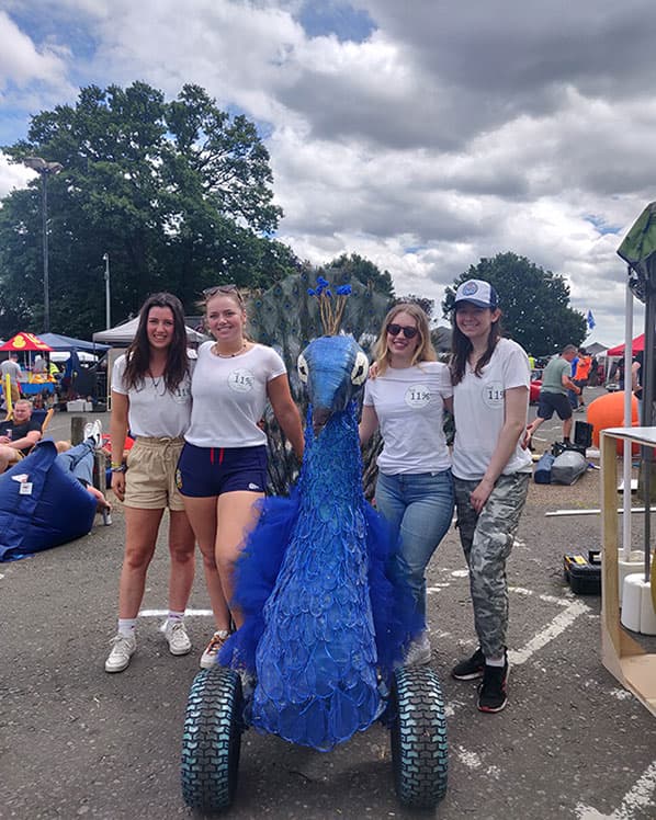 four students standing next to their RedBull soapbox which is decorated like a peacock