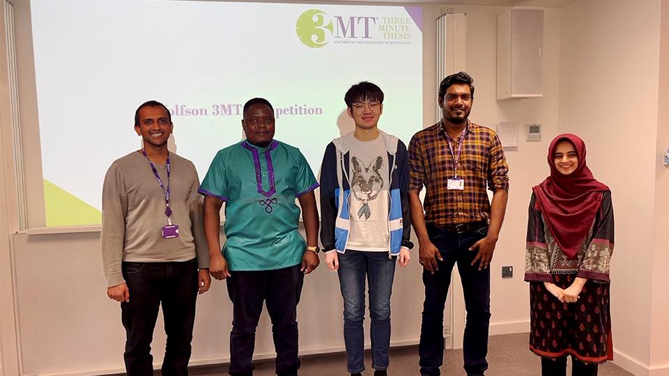 Five doctoral researchers are standing in a line in front of an interactive screen which reads 3MT Three Minute Thesis.