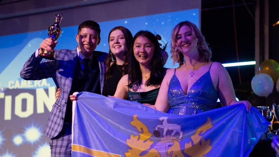 A group of male and female students standing together on a stage holding a David Collett hall flag, the students are smiling and representing their hall at an awards event.