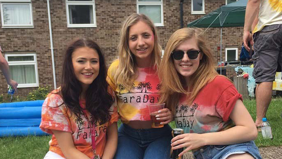 Three female students sitting outside on the grass outside Faraday hall. They are wearing brightly coloured Faraday hall t-shirts and smilling.