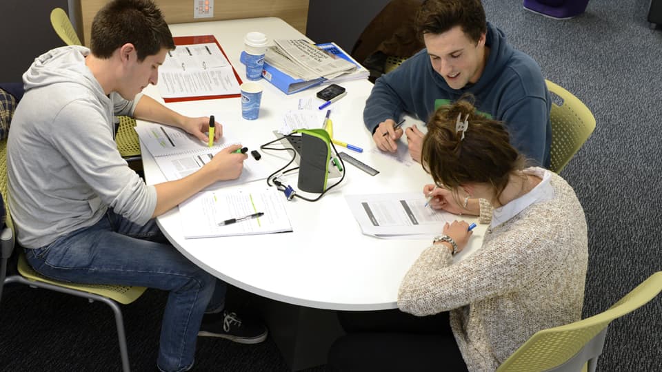 Two male students and one female tutor studying at a table with papers and pens
