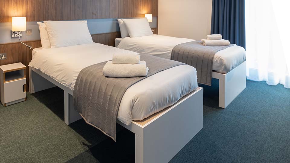 A twin bedroom in the Elite Athlete Centre: modern dark grey carpets and curtains, backed by a dark wood Scandi-style panelling, with a white bed covered in white bed linen, a grey bed topper with a couple of fluffy white towels artfully presented on the topper.