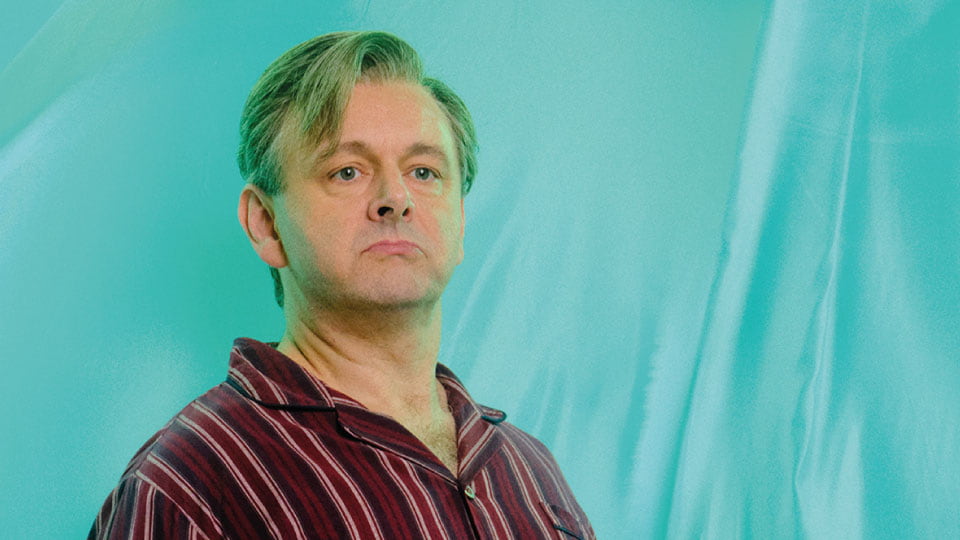A man with a serious look on his face, wearing a striped shirt in front of a blue fabric background.