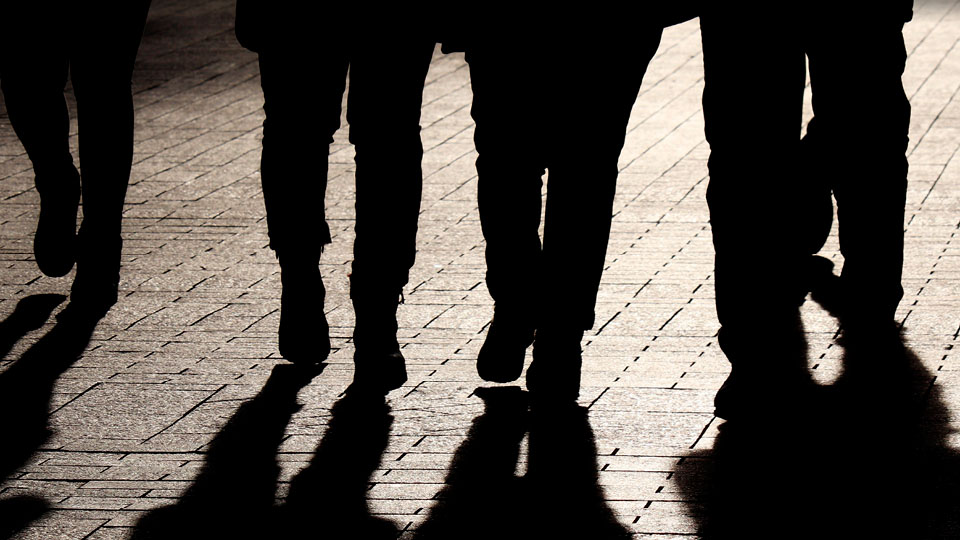 Black and white shadowy photograph of the lower half of four figures walking on a street