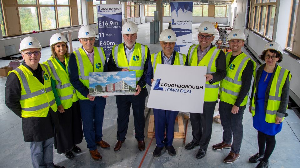 Eight people wearing hi-vis jackets and safety helmets, gathered in a new office space, launching the Loughborough Town Deal