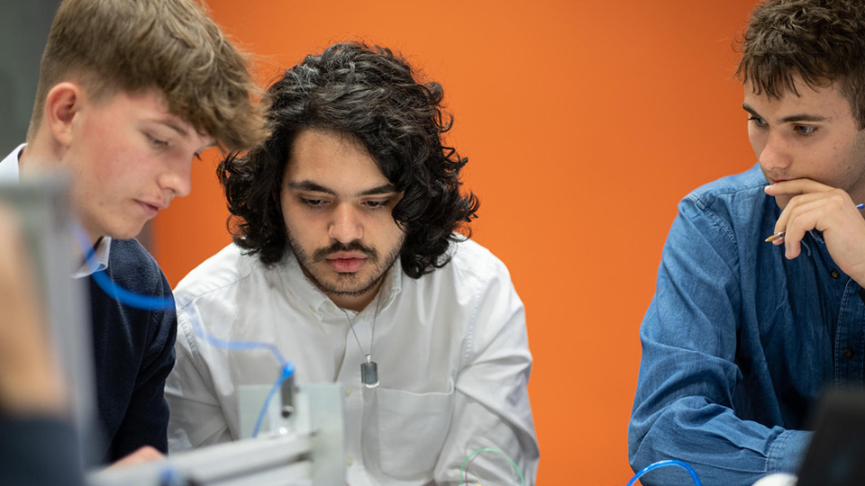 Photo of three male students looking at something together in an orange room