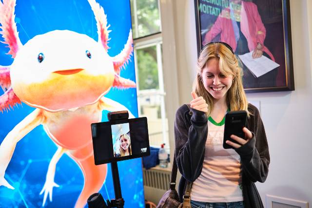 A woman smiling and looking at her phone, which is connected to an assistive robot. A picture of an axolotl can be seen in the background. 
