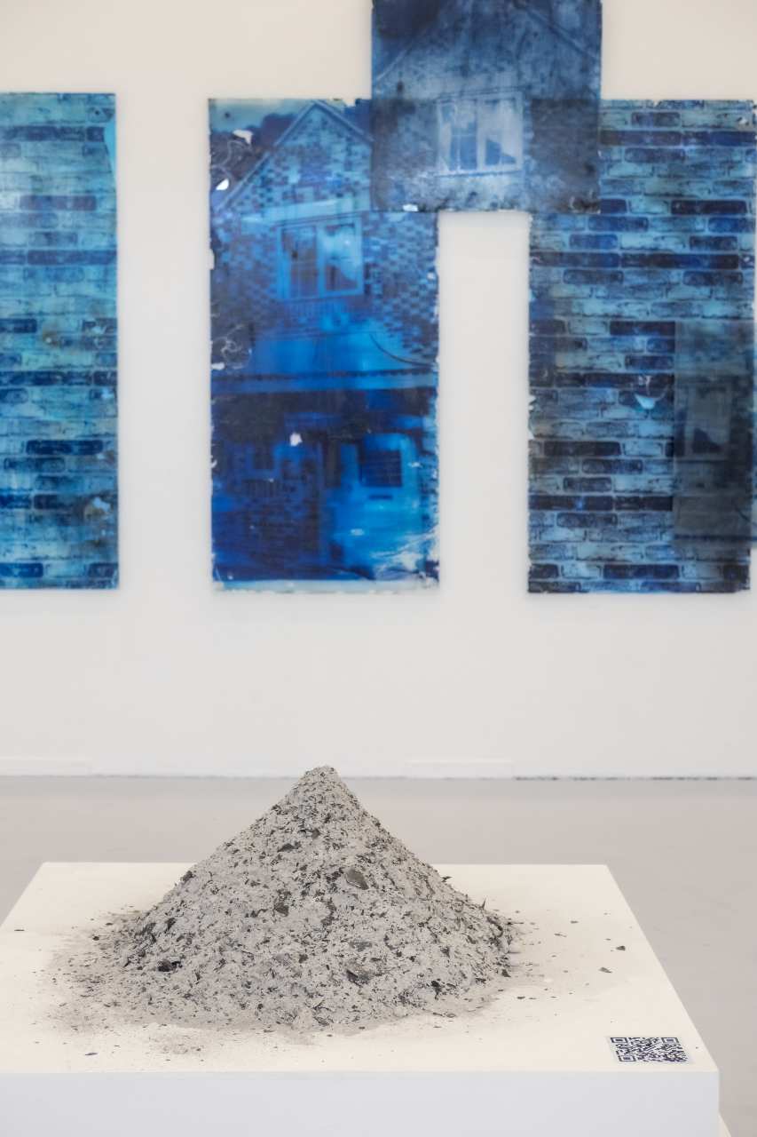 Pile of grey sand on top of a white plinth in a gallery, the wall in the background has four blue images of brick buildings.