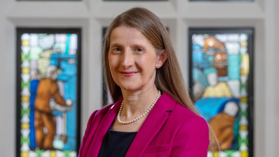 Headshot of Professor Rachel Thomson stood in front of stained glass windows wearing a pink blazer and black top
