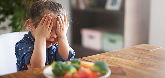 Pictured is a child with her hands on her face and a plate of vegetables. 