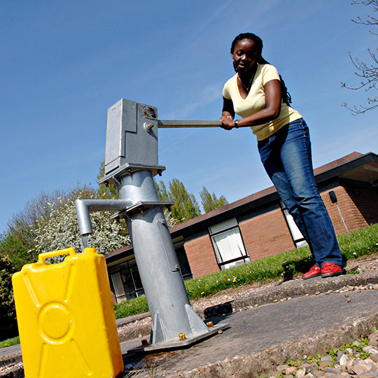 A woman using a manual water pump filling a yellow container with water
