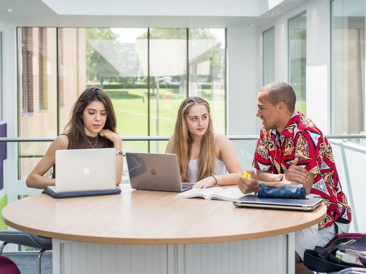 Three students (two female, one male) sitting around a table discussing their work. Two laptops are open in front of them.