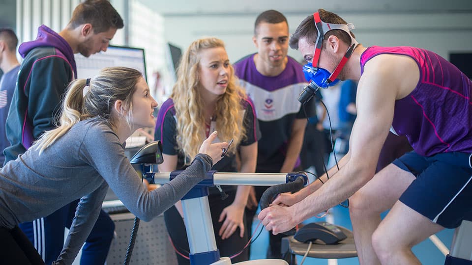 Sports science academics and students monitor an athlete wearing a face mask on an exercise bike 