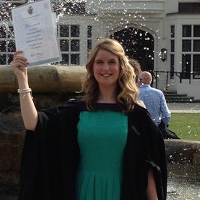 Amy Ward smiling at her graduation wearing the gown and holding the cap in front of the fountain