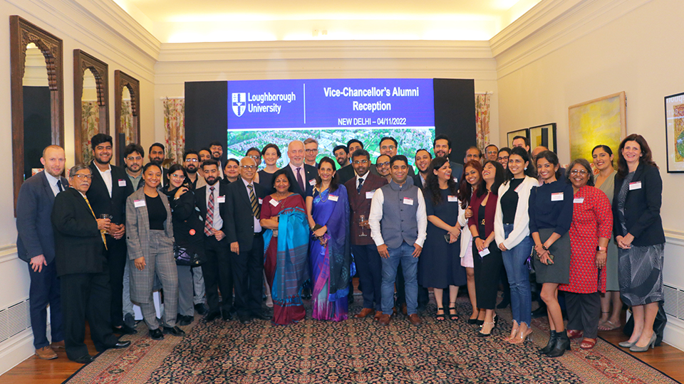 A group photograph of the event in New Delhi. Alumni and friends stand together in a large ornate room in front of a projector screen. 
