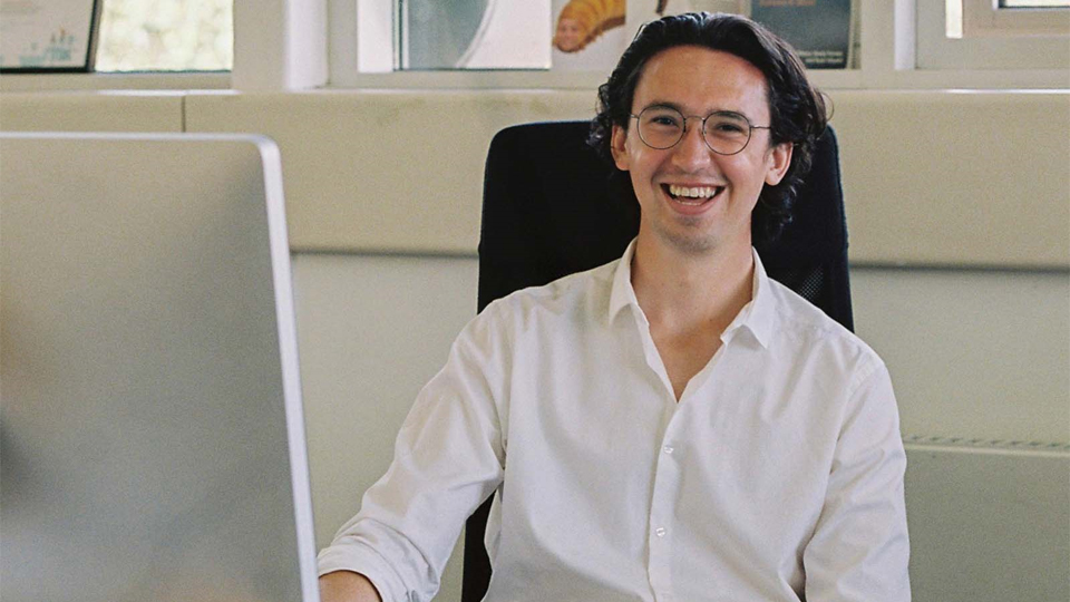 An image of Thomas Constant smiling whilst sitting a desk