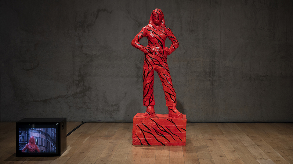 'Fetish or The Woman Who Built Her Own Statue' - a large red coloured statue of a woman is in a dark room and a tv screen is in the bottom left of the image
