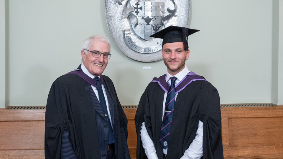 Murray and Ryan stand wearing gowns. In the background is a wooden panel and a silver University crest on the wall.