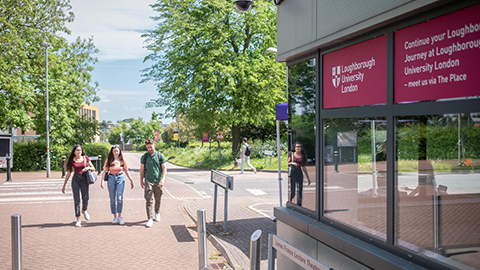 Three students walking on campus. A  building is on the right of the image and a road in view behind the students. 