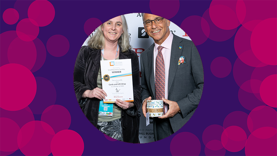 A purple background with pink circles on with an image of Catherine smiling in and holding her certificate next to Theo Paphitis who is holding her tape the middle of the canvas.