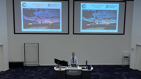 Simon Place stands in the centre of the lecture hall behind a lectern. There are two screens behind him with presentation slides displayed.