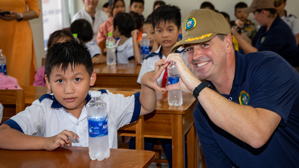 Capt Dransfield meets pupils from Hoa Dinh Tay School in Phu Yen, Vietnam. He is making a heart shape with his hand and the hand of a child. The children are seated at desks.