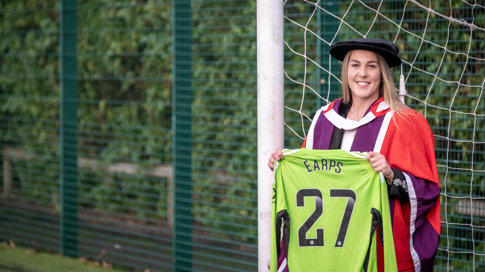 Mary Earps wearing a red gown and cap. She is standing in a football goal holding a green football jersey with her name printed on