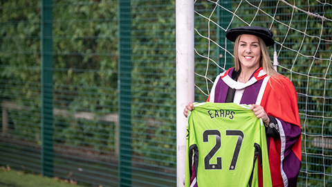 Mary Earps wearing a red gown and cap. She is standing in a football goal holding a green football jersey with her name printed on
