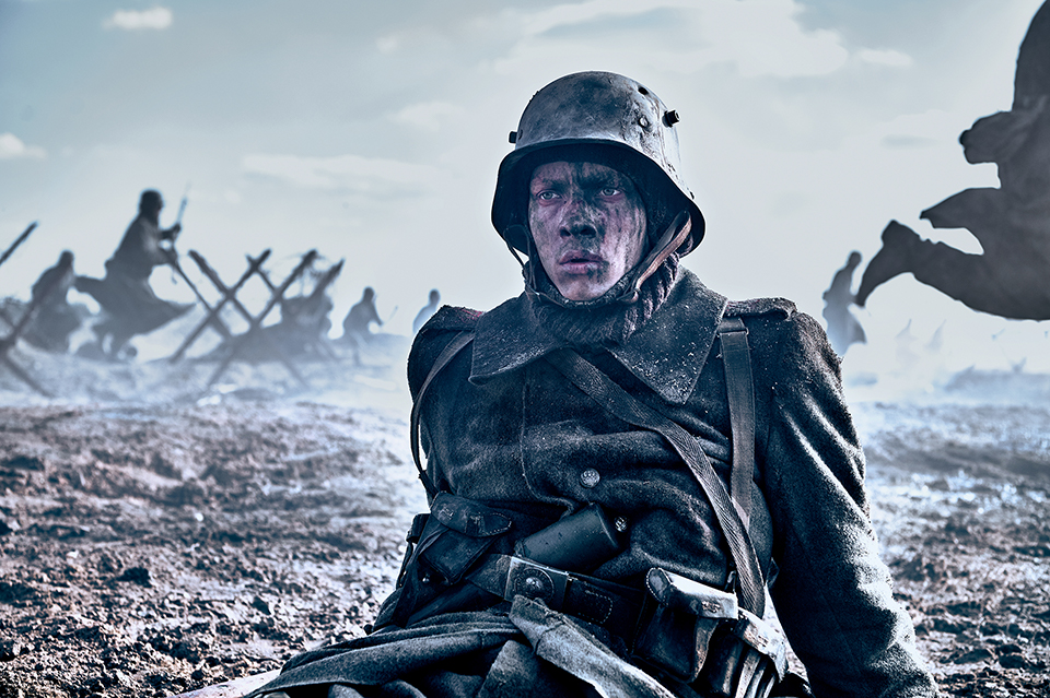 A dark image of a soldier sitting on the ground wearing uniform. His face is dirty. In the background is a war scene. Still taken from Netflix film All Quiet on the Western Front. Copywrite - ReinerBajo - Netflix.