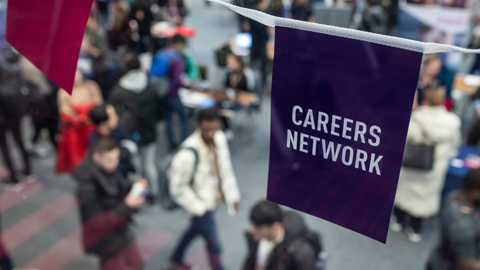 A blurred image of people walking in a room. There is a piece of bunting in purple that is in focus with the words "Careers Network" written on