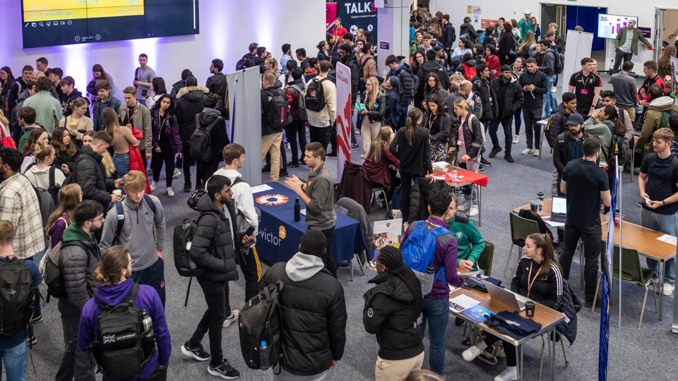 A room full of people meeting employers at exhibition stands. There are banners and tables. Many people are standing talking whilst others are walking around the space.