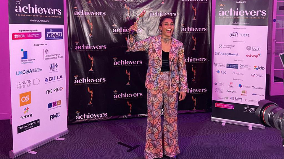 Aditi stands in front of a black backdrop at the awards evening. The backdrop has the achievers logos on. There are also two banners detailing awards sponsors. Aditi is wearing a colourful patterned suit and holds a trophy in her right hand as she smiles for the camera, which is pictured in the right of the image.