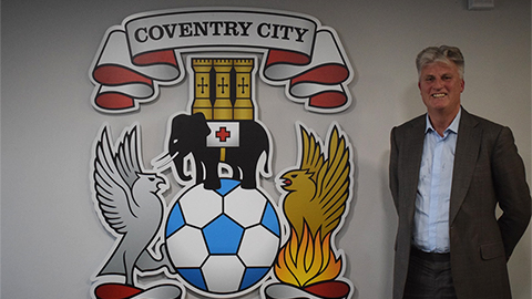 Doug King stands next to a Coventry City Football Club logo mounted on a wall