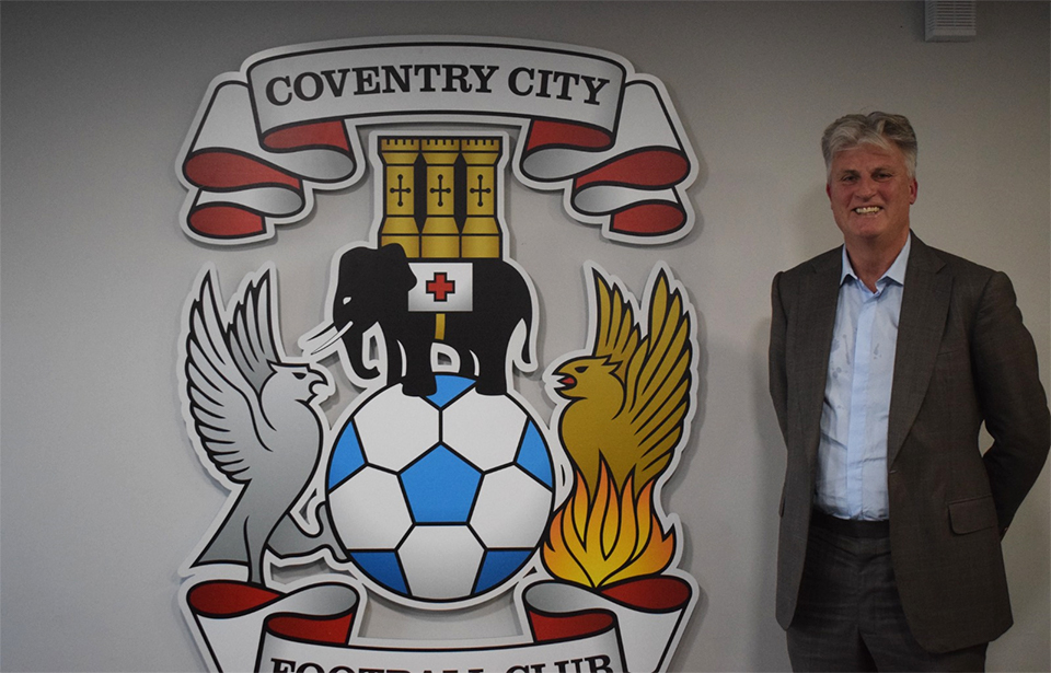 Doug King stands next to a Coventry City Football Club logo mounted on a wall
