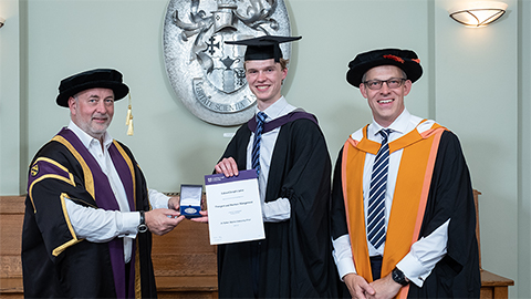 Three people pictured wearing graduation caps and gowns. Ed is in the middle holding a certificate and medal.