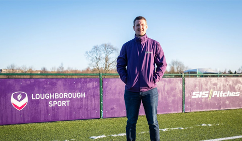 Joe stands on a pitch in front of purple signs. The signs have the Loughborough Sport and SIS Pitches logos on them. The image is taken on a clear day in January. There are some trees in the background.